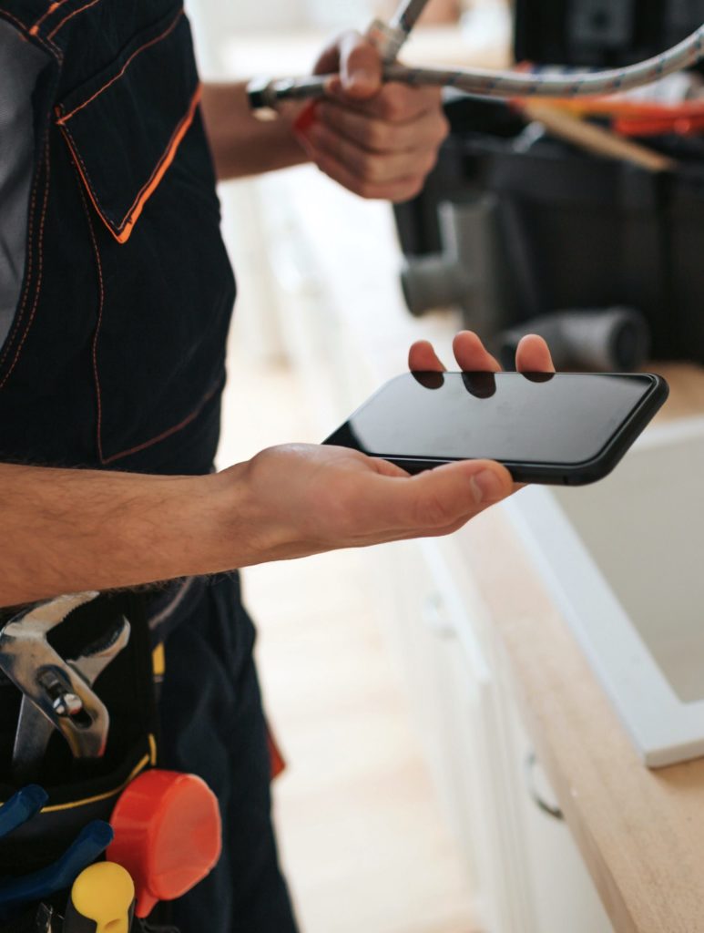 Cut view of man standing in kitchen at sink. He hold phone and wrench. Hose on desk.