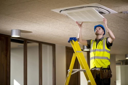 maintenance worker reinstalling ventilation panel on ceiling while standing on a ladder